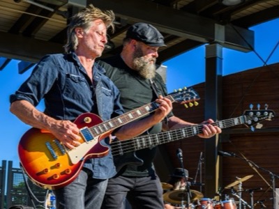 Two musicians perform outdoors with electric guitars under a covered structure against a blue sky, showcasing professional concert audio setup.