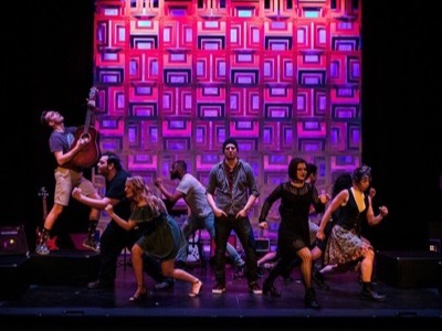 A live band performs on stage with vibrant purple and pink neon geometric backdrop lighting, demonstrating professional concert audio and lighting production.