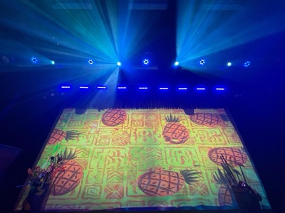 Colorful projection mapping displays vibrant geometric patterns and tropical fruit designs on a floor-mounted screen beneath blue stage lighting rigged overhead.