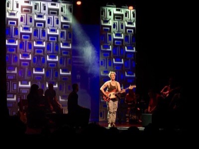 Musician performing on stage with guitar under concert lighting, backed by illuminated geometric panel displays and silhouetted audience members in foreground.