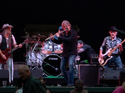 Live band performs on stage with professional audio equipment, including amplifiers and drums, while audience members watch from the foreground in a concert venue setting.