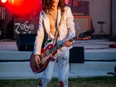 A guitarist performs on stage with a red semi-hollow body electric guitar, wearing a light-colored blazer and jeans, illuminated by red stage lighting at an outdoor evening event.