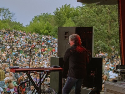 Musician performs at outdoor concert with keyboard, facing large crowd seated on hillside with lush green trees under clear sky.