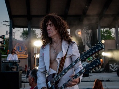 A long-haired guitarist performs on stage under professional lighting at an outdoor venue, with amplification equipment and audience visible in the background.