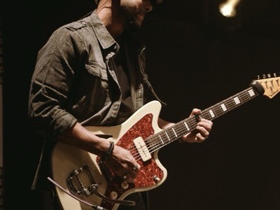 Guitarist performing on stage with professional concert lighting, playing a red and white electric guitar against a dark background.