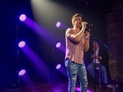 Male vocalist performing on stage with professional purple and white stage lighting from multiple overhead fixtures in a concert venue setting.