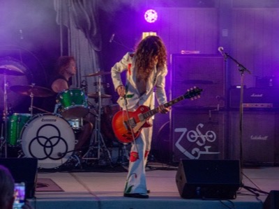 A musician performs on stage with a red guitar under purple lighting, with a drummer visible in the background and the band's logo displayed on the drum kit.