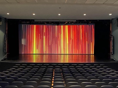 Interior view of a theater with rows of black seats facing a large projected video screen displaying vibrant red and yellow vertical light patterns.