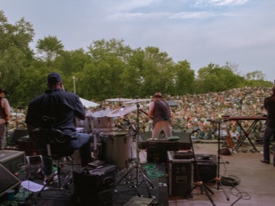 Musicians perform on an outdoor stage with professional audio equipment, addressing a large crowd seated in an amphitheater-style venue surrounded by trees.