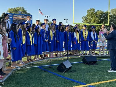 Graduation ceremony with students in blue robes and gold stoles standing on a field with professional sound equipment and microphones set up for the event.