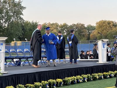 Graduation ceremony on outdoor stage with blue chairs, featuring graduates in caps and gowns with school officials, professional audio-visual staging, and audience seating.