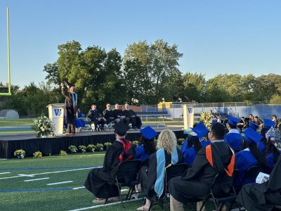 Outdoor graduation ceremony with a blue stage setup, podium with speakers, seated graduates in blue robes facing the stage, and trees lining the athletic field venue.