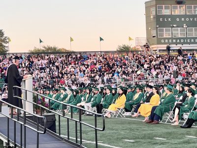 Large outdoor graduation ceremony with hundreds of students in green and gold regalia seated on a field, with a speaker at a podium and professional sound system setup visible.