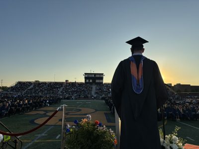 Graduate in cap and gown stands on outdoor stadium stage at sunset with crowded bleachers visible in background.