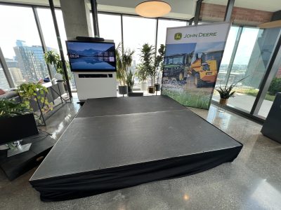 Black stage platform with riser setup in a modern office space with floor-to-ceiling windows, John Deere banner, display screen, and plants.